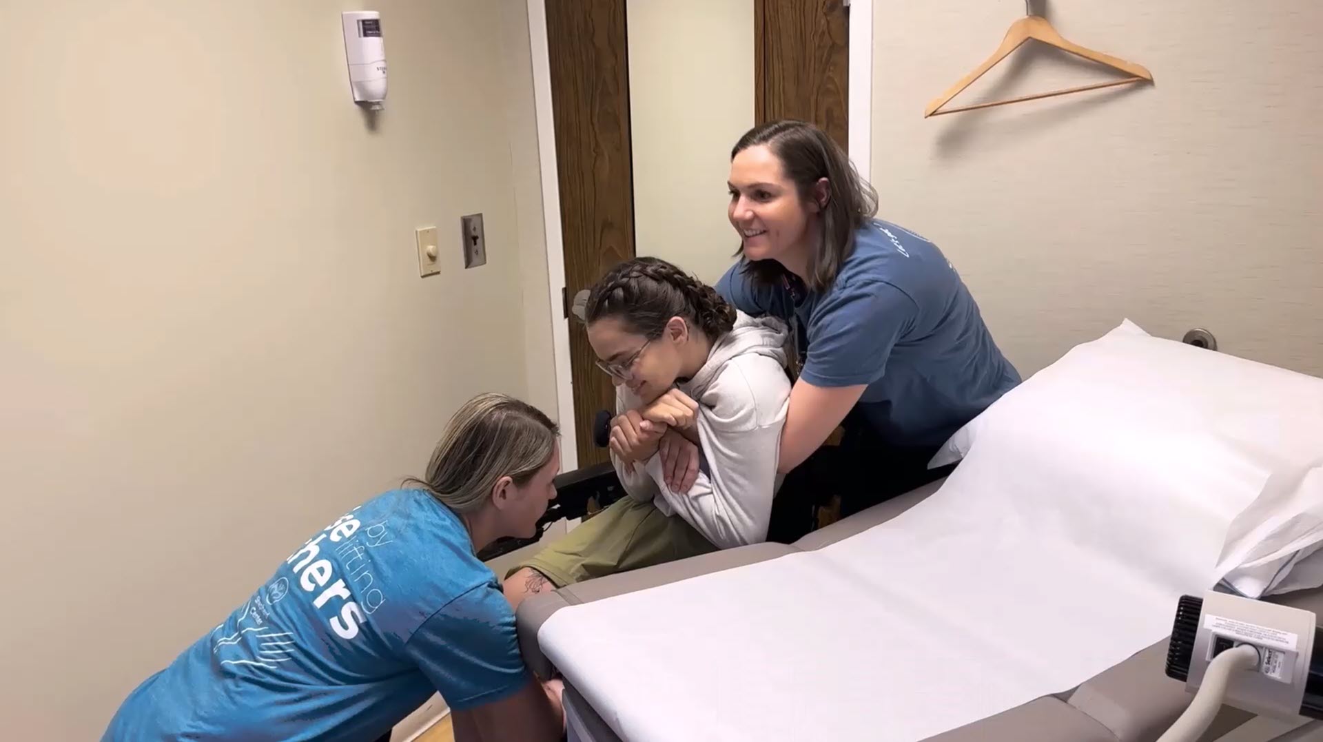 Two women assist another woman in transferring from an examination table in a medical room. The woman being helped is smiling and holding onto one assistant’s arms while the other supports her legs.