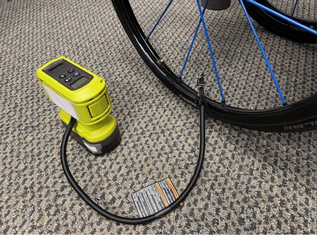A portable electric air pump is connected to a wheelchair tire and is resting on a carpeted floor.
