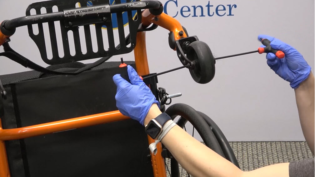A person wearing blue gloves uses tools to remove the front caster of an orange wheelchair.