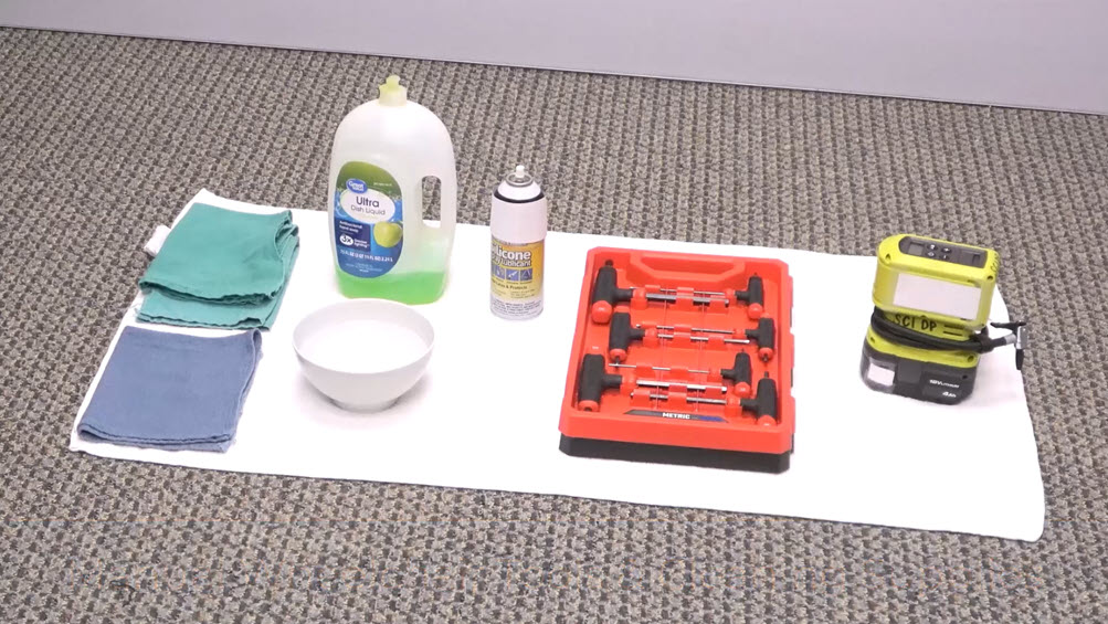 A white towel on a carpeted floor holds cloths, a bowl, a jug of dish soap, a can of silicone spray, a red set of wrenches, and a portable air pump, all neatly arranged in a row.
