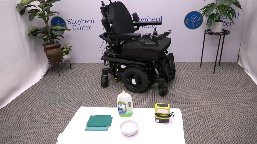 A motorized wheelchair sits against a wall with the Shepherd Center logo. In the foreground, a table holds a cloth, disinfectant, a bowl, and a yellow device. Potted plants are on either side of the wheelchair.