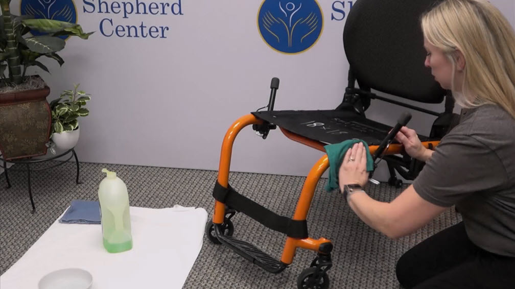 A woman cleans an orange wheelchair seat with a green cloth in a brightly lit room. A white towel, cleaning solution, and bowl are on the floor nearby. A Shepherd Center sign is visible on the wall behind her.