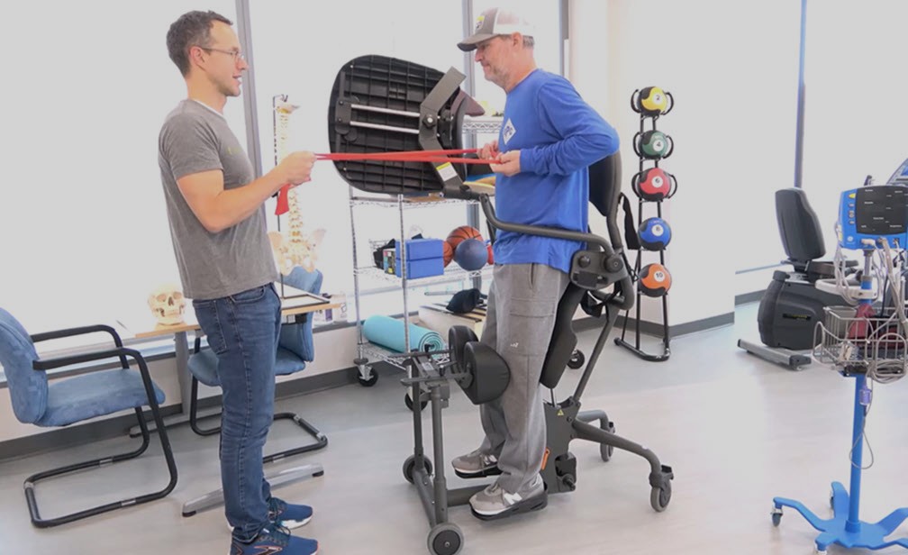 Two men are in a physical therapy setting; one stands while holding a red resistance band, the other, supported by a standing frame, pulls the resistance band. Therapy equipment and a skeleton model are in the background.