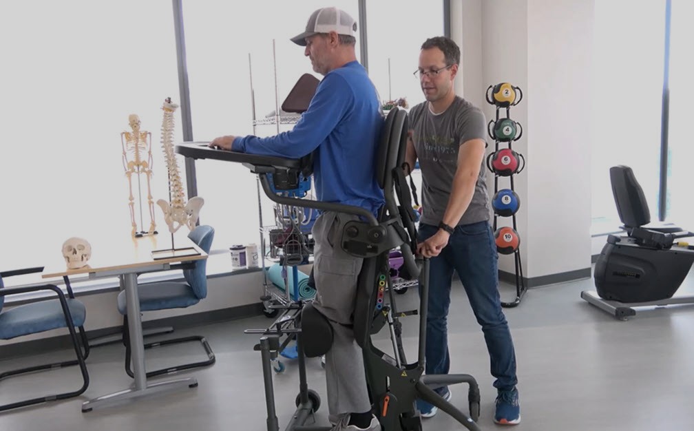 A man uses a standing frame with support while another man assists him in a bright physical therapy room with anatomical models and exercise equipment in the background.