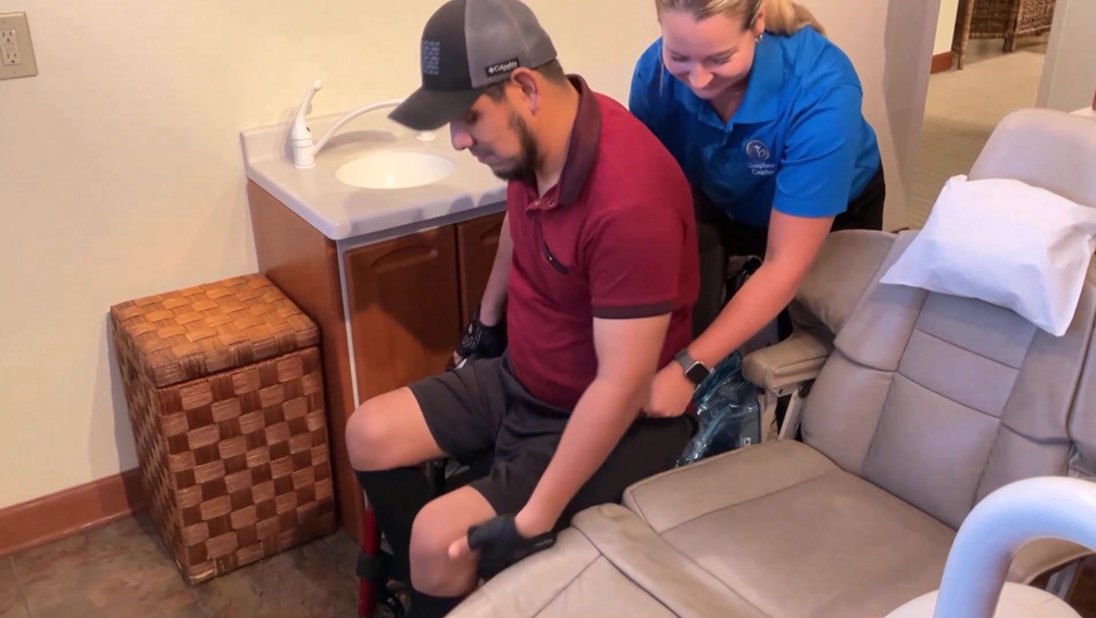 A woman in a blue shirt assists a man in a maroon shirt and gray cap as he transfers from a wheelchair to a recliner chair in a room with a sink and wicker basket.