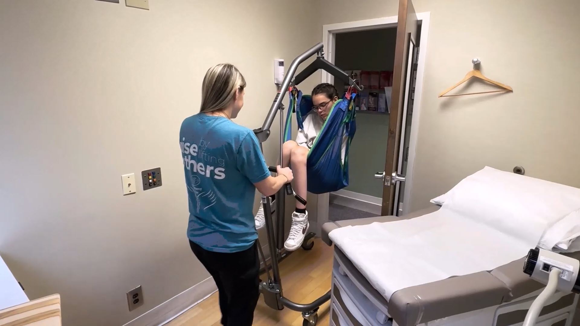 A caregiver uses a Hoyer lift to help a patient transfer from a wheelchair to an exam table in a clinic room.