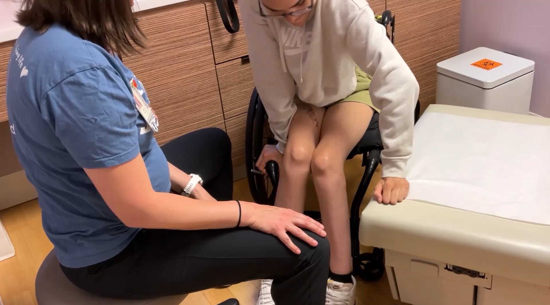 A person in a wheelchair prepares to transfer onto a medical exam table with assistance from a healthcare worker sitting nearby.