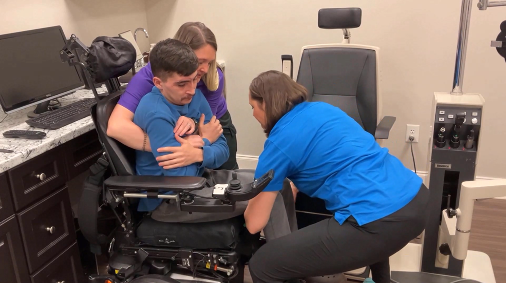 Two women assist a young man in a blue shirt as he is transferred from a power wheelchair to a medical exam chair in a clinical setting. Medical equipment and a computer are visible in the room.