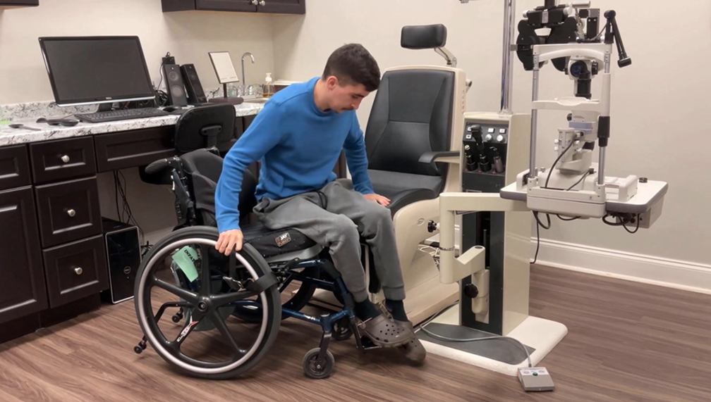 A man in a blue sweater and gray pants transfers from a wheelchair to an exam chair in an eye doctor's office, with medical equipment and a computer visible in the background.