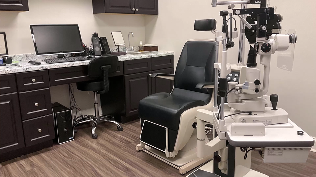 An eye exam room with a black exam chair, a slit lamp machine, a desk with a computer, office chair, cabinets, and a sink, all on a wood floor.