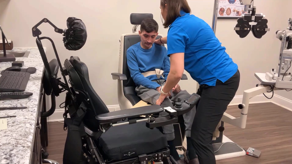 A man in a blue shirt sits in an eye exam chair while a woman in a blue uniform helps adjust his position. An empty wheelchair is next to him, with medical equipment visible in the room.