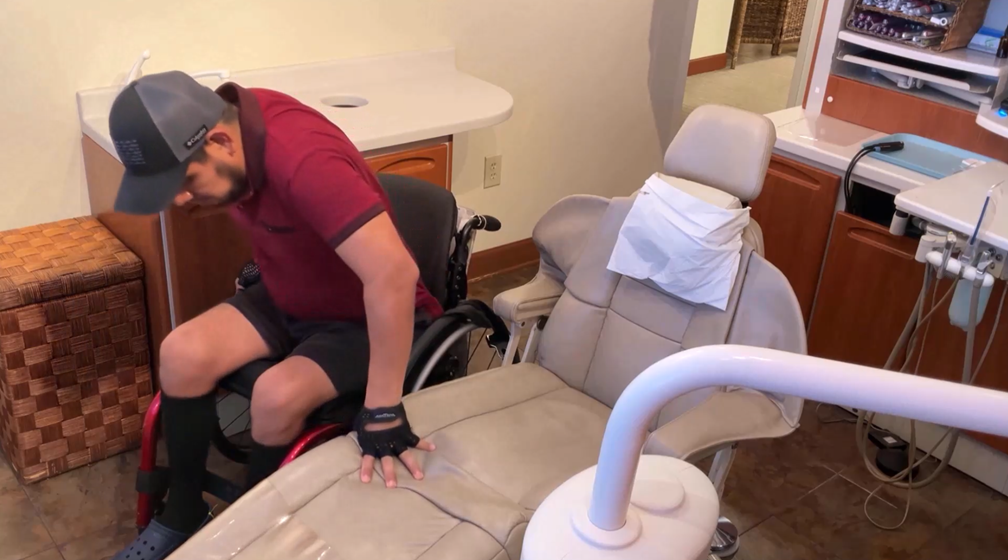 A man in a wheelchair transfers himself onto a dentist chair in a dental clinic. He is wearing a maroon shirt, shorts, a cap, and gloves, with his left hand on the dentist chair for support.