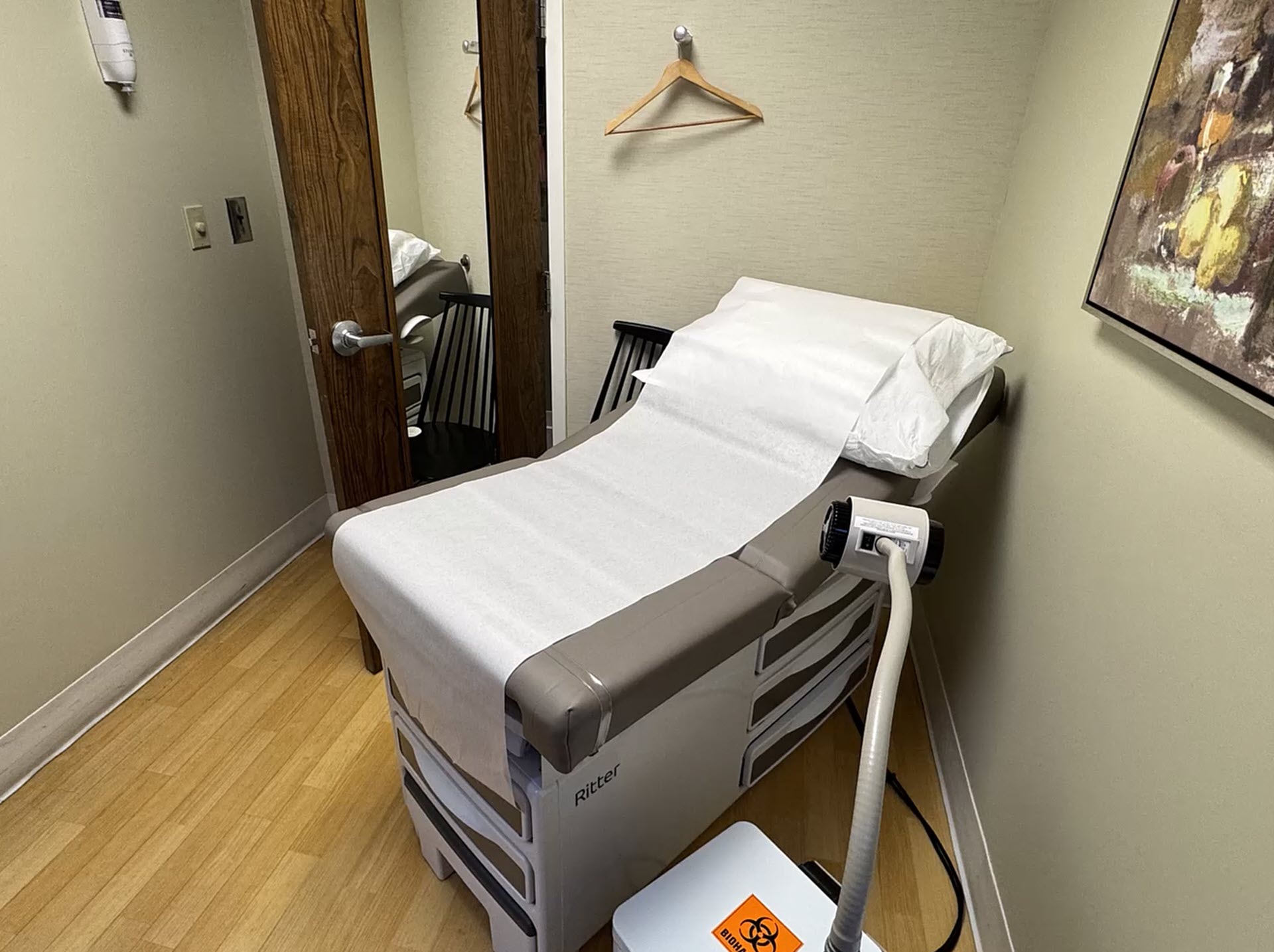 A small medical exam room with an exam table, pillow, and paper covering, a wooden chair, a hanger on the wall, a mirror on the door, and a medical device with a biohazard label on the floor.