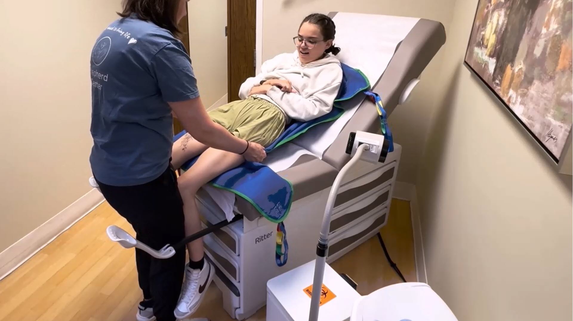 A healthcare provider assists a young woman lying on an exam table in a medical office. Medical equipment and artwork are visible in the room.