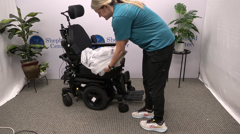 A person in a teal shirt and black pants places a clear plastic cover over the seat of a black power wheelchair, demonstrating daily living after neurological injury, in a room with plants and Shepherd Center logos on the walls.