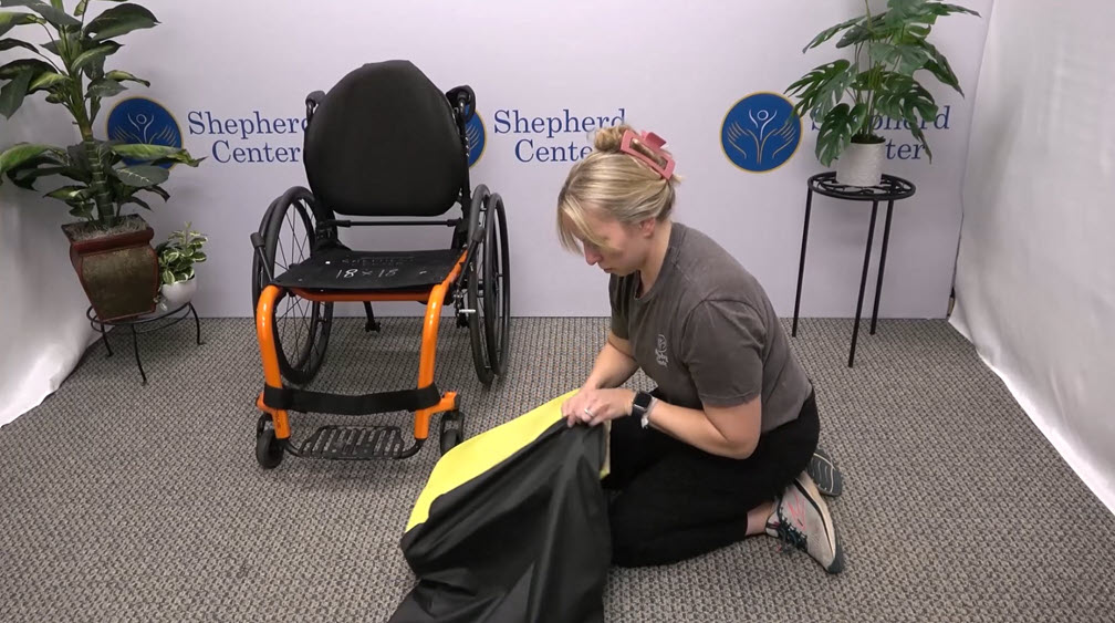 A woman kneels on the floor, putting a yellow cushion inside a black cloth cover. Behind her is an empty wheelchair with orange accents, a white backdrop with 