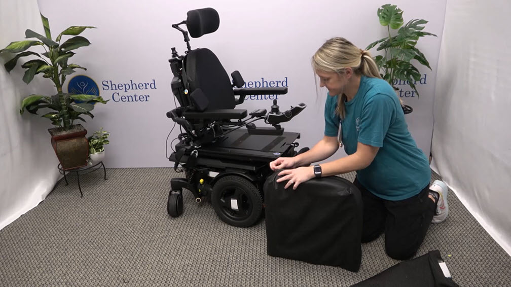 A woman kneels next to a black power wheelchair, attaching a black cover to the seat. The scene is indoors with potted plants and a Shepherd Center banner in the background.