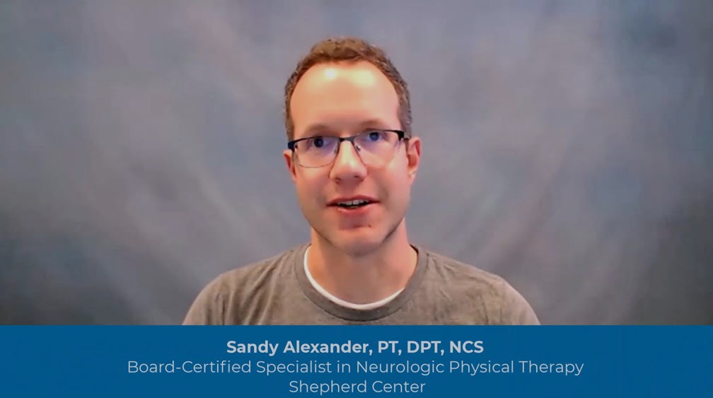 A man with short brown hair and glasses, wearing a gray T-shirt, speaks in front of a plain blue-gray background. Text below reads: 