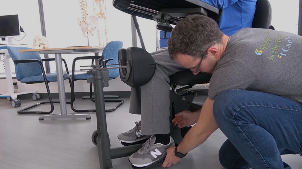 A man in a gray shirt adjusts the foot placement of another person seated in a standing frame in a clinical setting. There are blue chairs and anatomical models in the background.