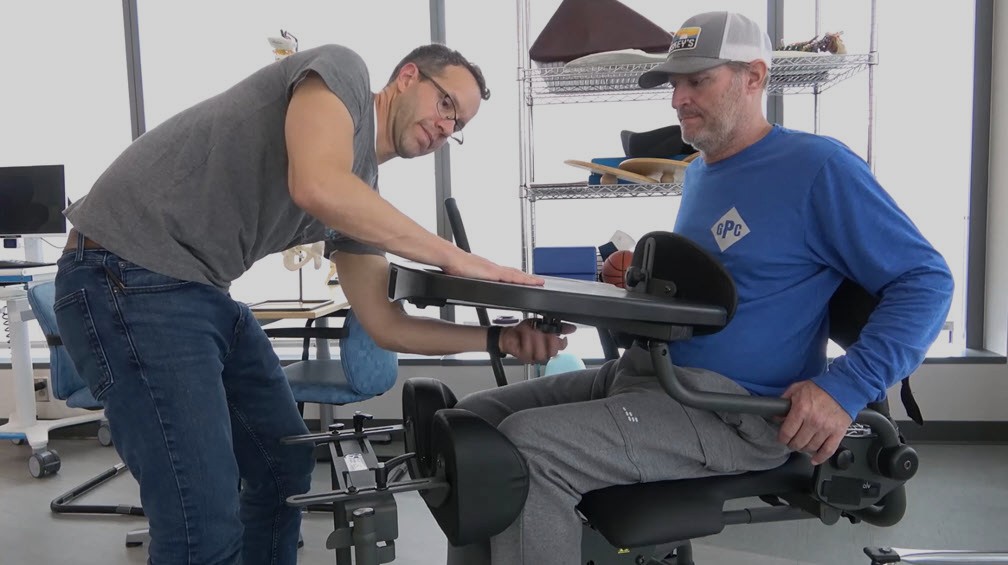 A man in a gray t-shirt adjusts a large black chest pad and tray table for a man seated in a standing frame, who is wearing a blue long-sleeve shirt and gray sweatpants, in a brightly lit therapy room with shelves and equipment in the background.