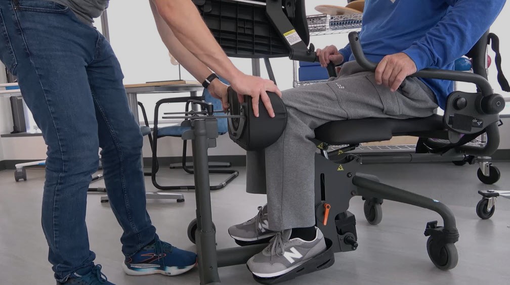A person sitting in a wheelchair places their lower leg into a padded knee brace as another person adjusts the device for physical therapy or rehabilitation purposes in a clinical setting.