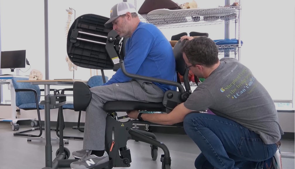 A man in gray sweatpants and a blue shirt sits in a standing frame while another man crouches beside him, making adjustments to the frame in a rehabilitation setting.