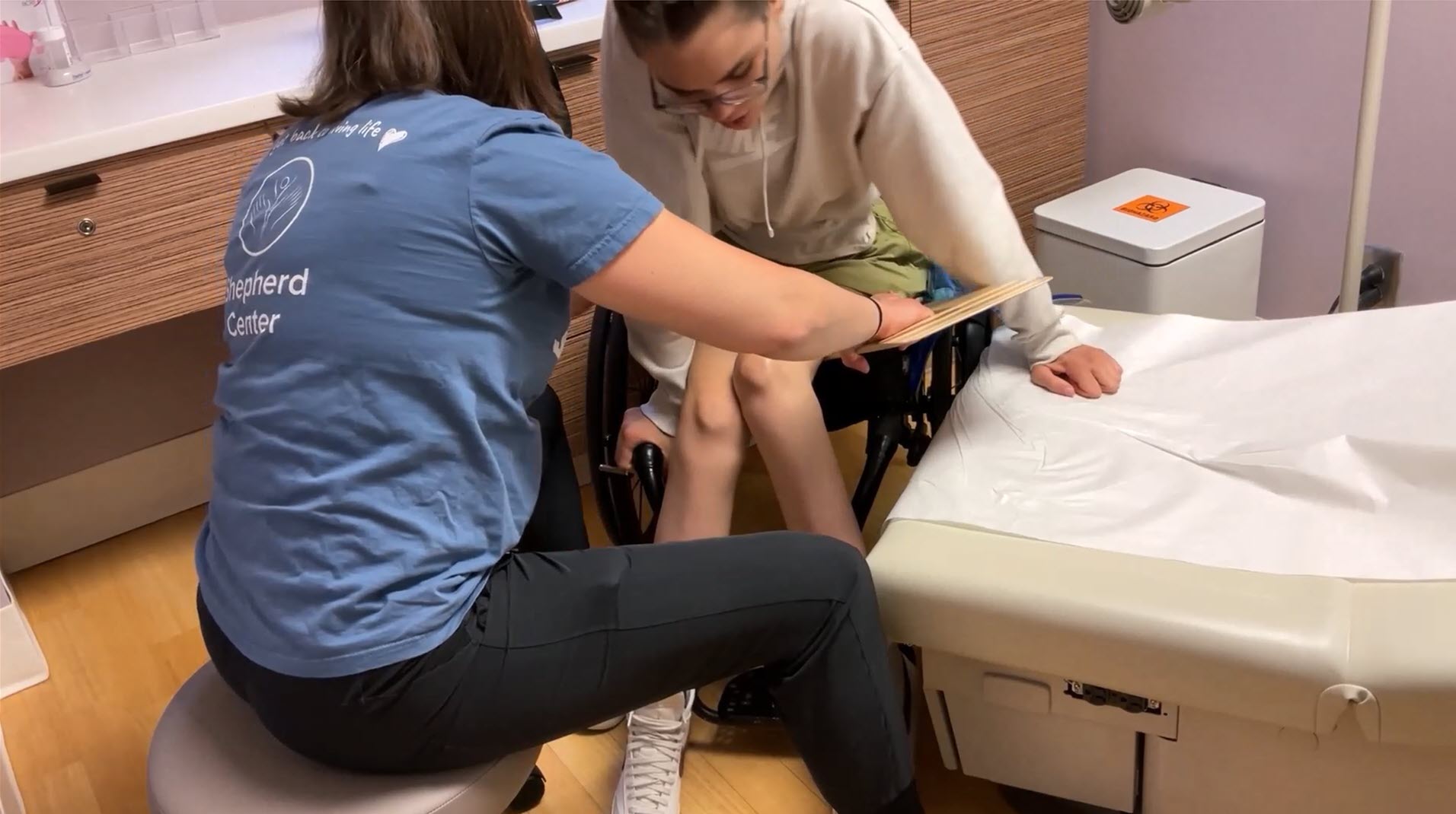 A person in a blue shirt assists another person, who is in a wheelchair, as they transfer onto a medical exam table in a clinical setting.