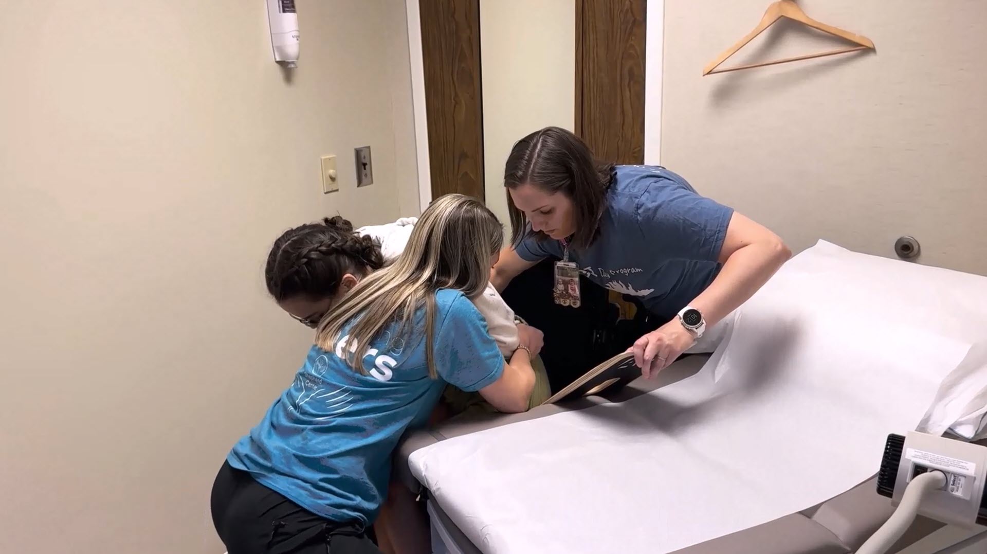 Two women assist another woman in a wheelchair in getting onto a medical exam table in a clinic room. One woman supports her from behind, while the other supports her from the front.
