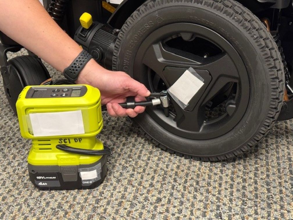 A person uses a portable electric air pump to inflate the rear tire of a wheelchair. The pump is connected to the tire’s valve, and the wheelchair is on a carpeted floor.