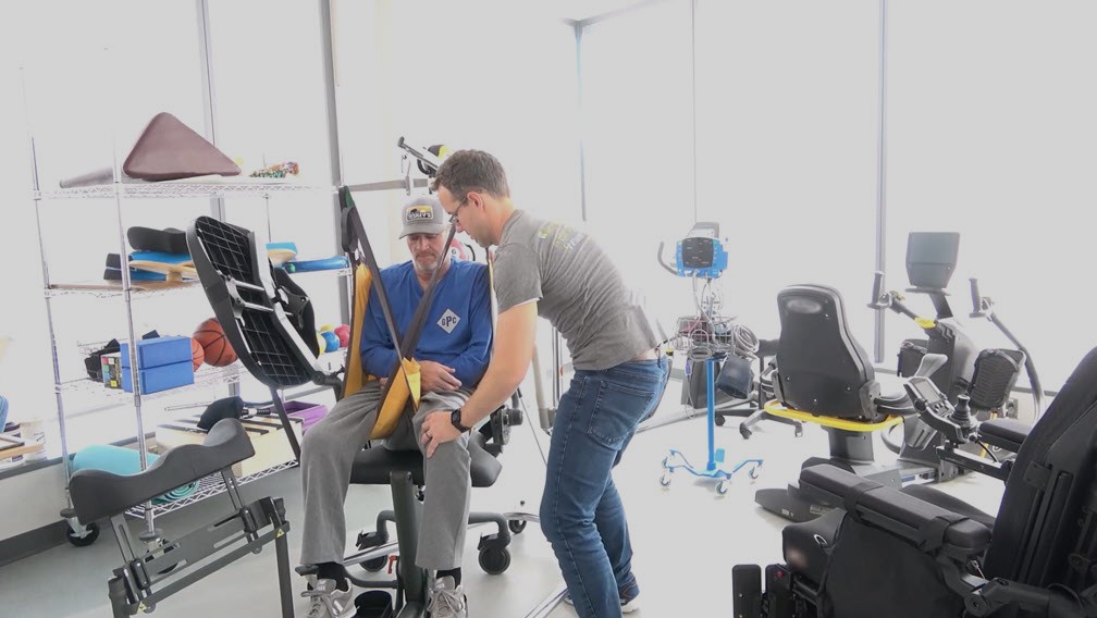 A man wearing a cap sits on a chair with a support sling, while another man assists him in a bright rehabilitation room focused on exercise and wellness after traumatic injury, with various equipment and wheelchairs visible.