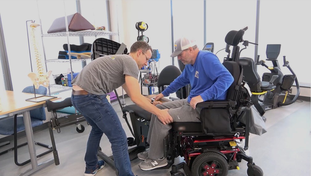 A man in a gray t-shirt helps another man wearing a blue shirt and cap transfer from a motorized wheelchair to a chair in a rehabilitation center with equipment in the background.
