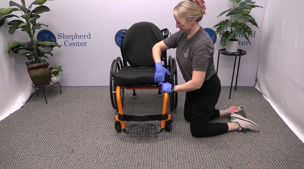 A woman wearing gloves kneels on the floor while preparing to clean a wheelchair in a room with plants and a Shepherd Center sign on the wall.