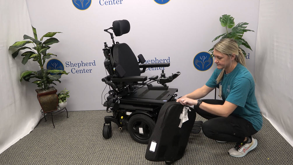 A person kneels beside a black power wheelchair, inspecting or attaching a component, in front of a Shepherd Center backdrop and potted plants.