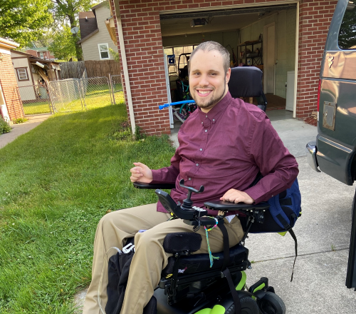 A smiling man in a maroon shirt and khaki pants sits in a green and black power wheelchair on a driveway outside a brick house, with grass and a fence nearby.