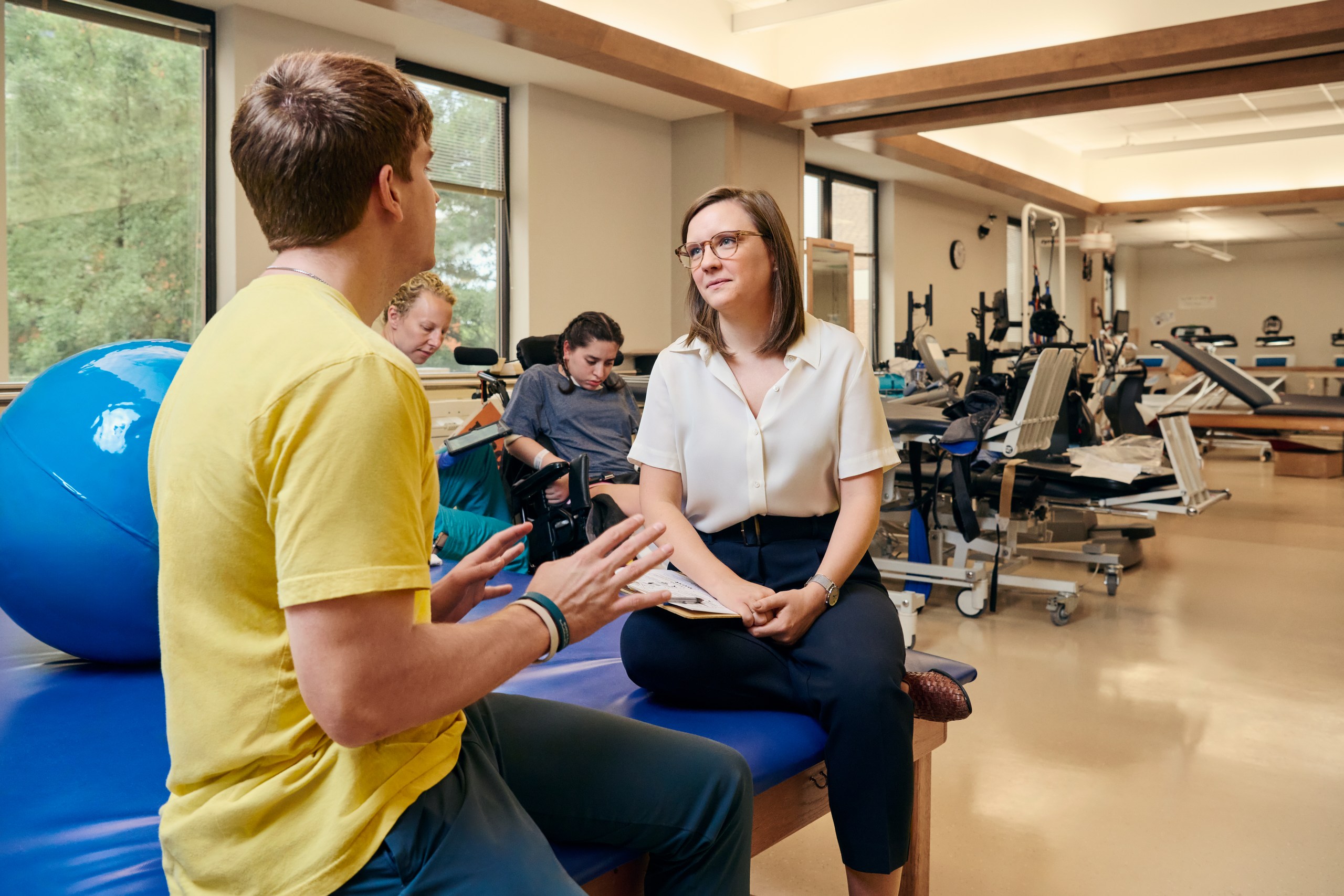 A person in a yellow shirt and a person in glasses converse intimately about brain injury while seated in a physiotherapy room filled with exercise equipment and a large blue exercise ball. Two others are focused on family training, engaged in rehabilitation exercises.