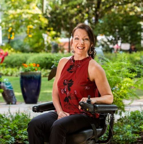 A smiling woman with brown hair, wearing a sleeveless red and black top and black pants, sits outdoors in a power wheelchair on a sunny day, surrounded by greenery and colorful plants.